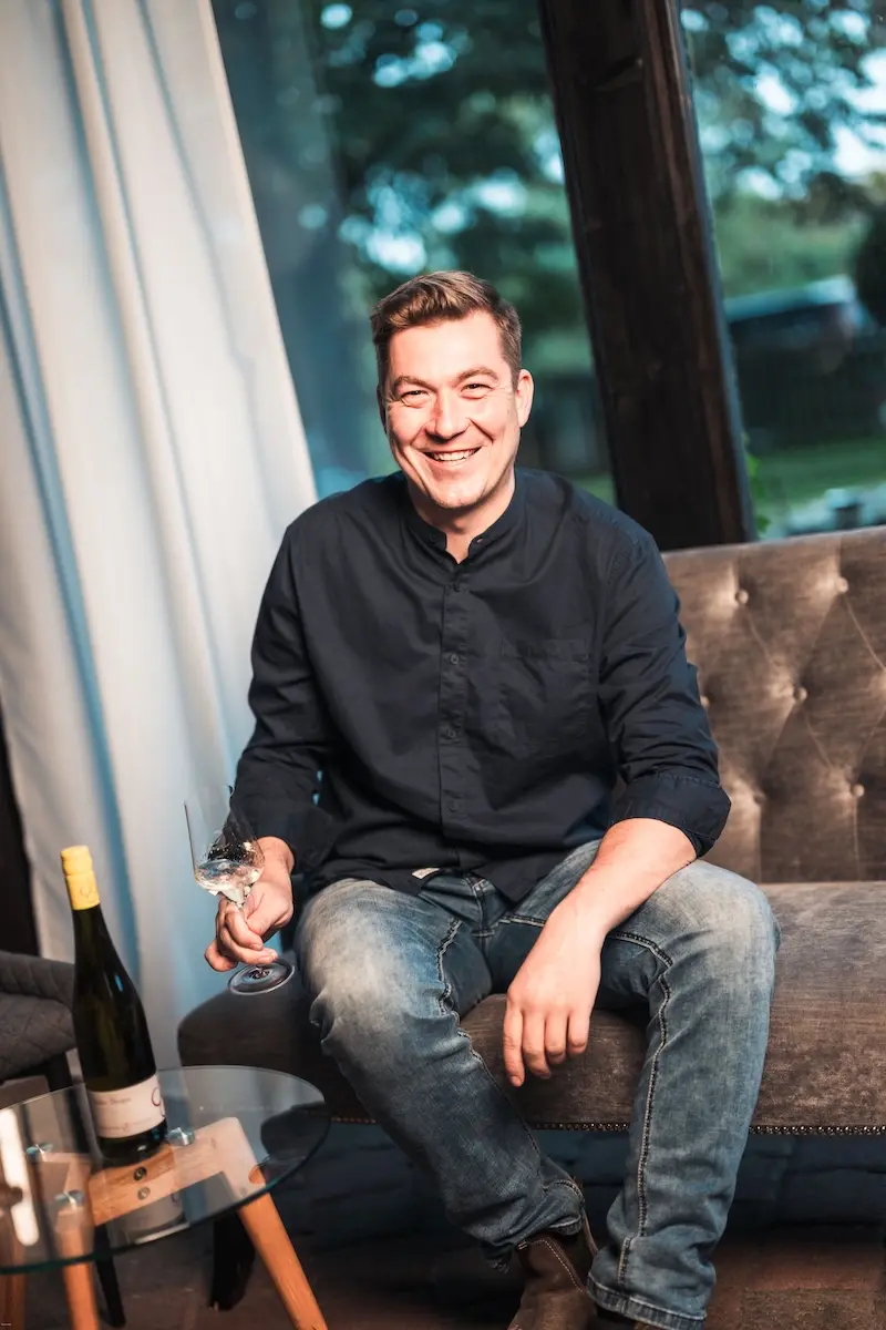 Johannes Zwicker sitzt lachend mit einem Glas Weißwein auf einem Sofa